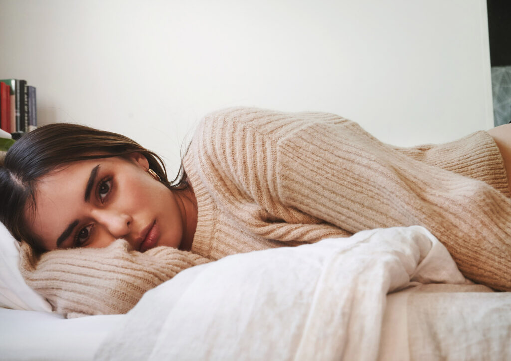 Marintia Bustos lies on a bed wearing a beige sweater with a contemplative expression surrounded by books and a plant.